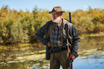 Senior man with gun going to hunt on wild ducks on lake. Male looking away