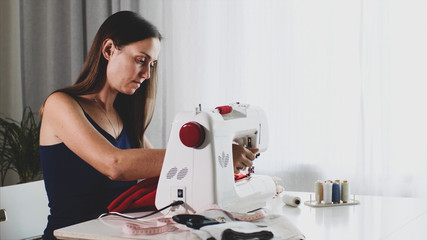 Young woman tailor is sewing red cloth on sewing machine. Seamstress is working at home sitting at...