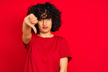 Fototapeta na wymiar Young arab woman with curly hair wearing casual t-shirt over isolated red background looking unhappy and angry showing rejection and negative with thumbs down gesture. Bad expression.