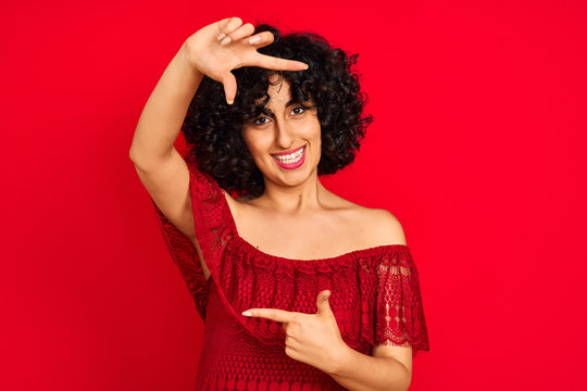 Young arab woman with curly hair wearing casual dress over isolated red background smiling making frame with hands and fingers with happy face. Creativity and photography concept.