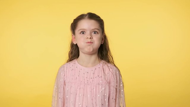 Funny little girl playfully making faces looking at camera. Positive emotion. Caucasian kid girl in pink dress doing silly funny face puffing out cheeks at yellow background. Medium shot in 4k, UHD
