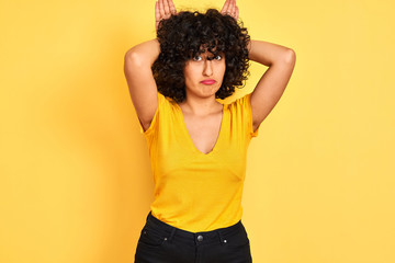 Young arab woman with curly hair wearing t-shirt standing over isolated yellow background Doing bunny ears gesture with hands palms looking cynical and skeptical. Easter rabbit concept.
