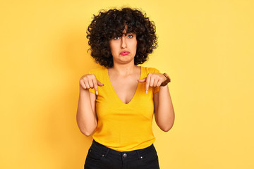 Young arab woman with curly hair wearing t-shirt standing over isolated yellow background Pointing down looking sad and upset, indicating direction with fingers, unhappy and depressed.