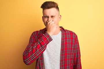 Young handsome man wearing casual shirt standing over isolated yellow background smelling something stinky and disgusting, intolerable smell, holding breath with fingers on nose. Bad smells concept.