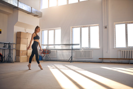 Cheerful Young Female Athlet, Dressed In Black Sportswear, Walking Gracefully On Tiptoe In Roomy Brightly Lighted Gym Hall, Spreads Hands Sideways, Looking Away, Professional Sport Concept