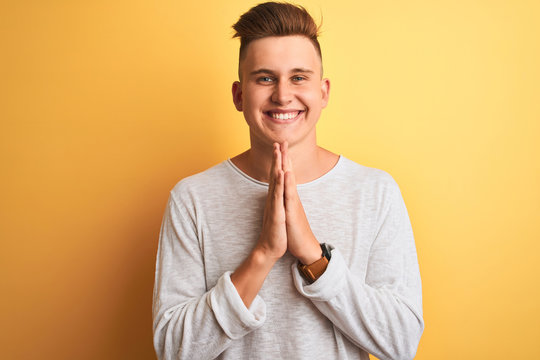 Young handsome man wearing white casual t-shirt standing over isolated yellow background praying with hands together asking for forgiveness smiling confident.