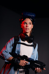 Young female holding weapon, looking side in search, special mask device on head. Black background