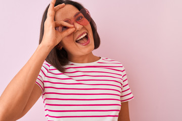 Young beautiful woman wearing fashion sunglasses with hearts over isolated pink background with happy face smiling doing ok sign with hand on eye looking through fingers