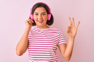 Young beautiful woman listening to music using headphones over isolated pink background doing ok sign with fingers, excellent symbol