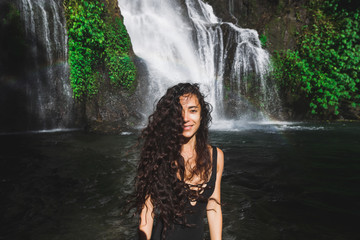 Young slim brunette woman with curly hair enjoying in lagoon of huge tropical waterfall Banyumala in Bali. Wearing in black swimsuit. Happy vacations in Indonesia. Wanderlust travel concept.