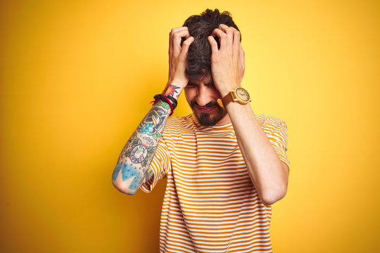 Young man with tattoo wearing striped t-shirt standing over isolated yellow background suffering from headache desperate and stressed because pain and migraine. Hands on head.
