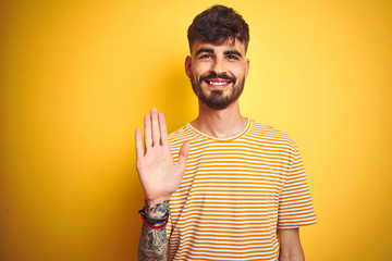 Young man with tattoo wearing striped t-shirt standing over isolated yellow background Waiving...