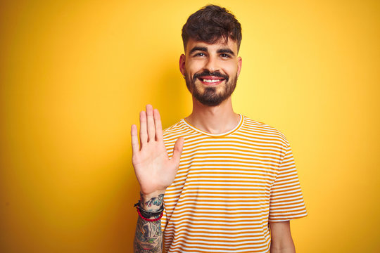 Young man with tattoo wearing striped t-shirt standing over isolated yellow background Waiving saying hello happy and smiling, friendly welcome gesture