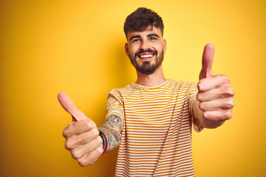 Young man with tattoo wearing striped t-shirt standing over isolated yellow background approving doing positive gesture with hand, thumbs up smiling and happy for success. Winner gesture.