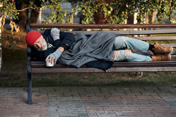 Vagrant male lying on street bench asking for money, for any help. Desperate and lonely homeless man without shelter