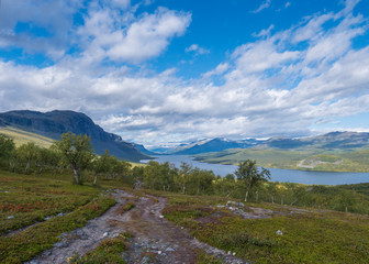 Fototapeta na wymiar Lapland landscape with beautiful river Lulealven, snow capped mountain, birch tree and footpath of Kungsleden hiking trail near Saltoluokta, north of Sweden wild nature. Summer blue sky