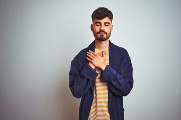 Young man with tattoo wearing bathrobe standing over isolated white background smiling with hands on chest with closed eyes and grateful gesture on face. Health concept.