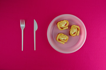 A few pieces of biscuit roll are in a white disposable plastic plate, and next to them are a fork and a knife.