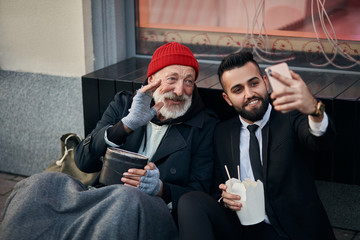 Genial rich man in costume making selfie with caucasian beggar sitting on street, happy