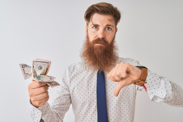 Young redhead irish businessman holding dollars standing over isolated white background with angry...