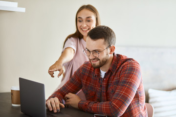 Cheerful beautiful young couple spending time in front of the computer, watching funny video, excited girl stretching hand towards screen pointing at device, positive atmosphere, portrait