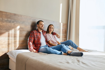 Portrait of charming lady casually dressed sitting happily with young unshaven man on big bed, on background of beautiful curtains and light window, man watching TV hugging on shoulders, family