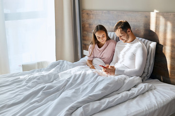 Obraz na płótnie Canvas Young husband and wife having honeymoon, texting to friends about arrival to other country, travelling together, resting in hotel room, using cell phone to message, love concept