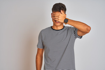 Young indian man wearing navy striped t-shirt standing over isolated white background smiling and laughing with hand on face covering eyes for surprise. Blind concept.