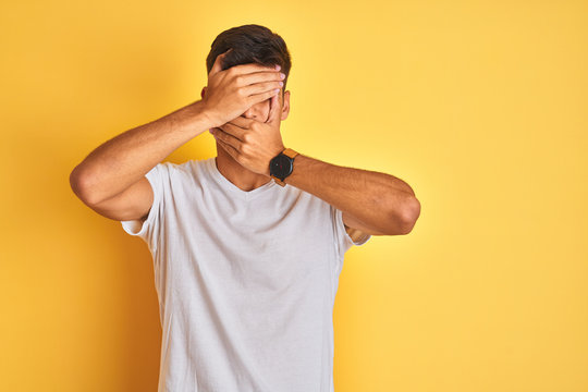 Young indian man wearing white t-shirt standing over isolated yellow background Covering eyes and mouth with hands, surprised and shocked. Hiding emotion