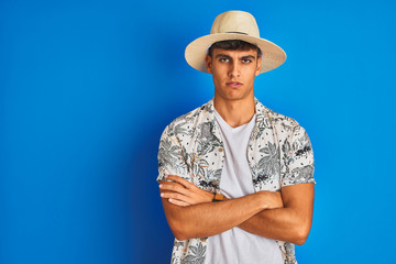 Indian man on vacation wearing hawiaian shirt summer hat over isolated blue background skeptic and nervous, disapproving expression on face with crossed arms. Negative person.