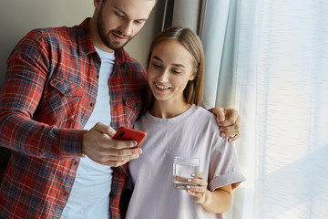 Cheerful young couple in the morning standing by bright window keeping glass of water and cell phones in hands, watching funny video at home, man hugging gently, family portrait
