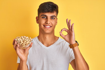 Young indian man holding bowl with pistachios over isolated yellow background doing ok sign with fingers, excellent symbol