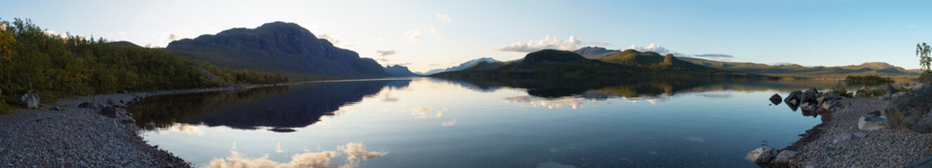 Wide panoramic view on river Lulealven in Saltoluokta in Sweden Lapland during sunset golden hour. Green mountain, birch trees, rock boulders clouds and sky reflecting at clear the water.