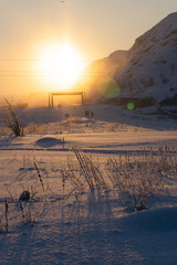 the bright orange sun at dawn in the north beyond the Arctic Circle illuminates the small grass, pipes and old rural houses on a frosty morning in winter
