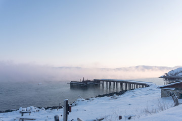 old wooden pier for large ships on the river bank covered in thick white fog from the water at dawn on a frosty winter day outside the Arctic Circle