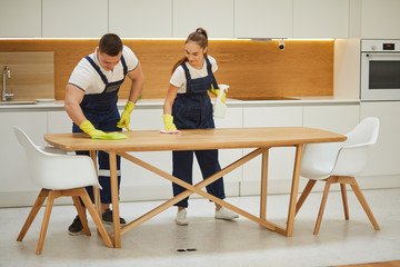 Obraz premium Teamwork of cleaning staff in kitchen with white interior. Preparing kitchen for check in. Man rubs the table with rag, woman help him