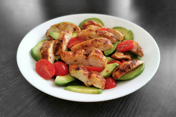 Fototapeta na wymiar Grilled chicken salad with avocado and tomatoes on a white plate with black gray wood table background.