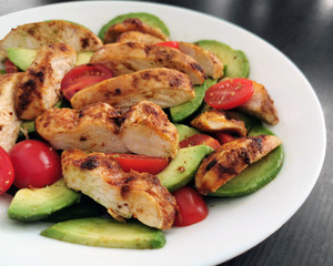 Grilled chicken salad with avocado and tomatoes on a white plate with black gray wood table background.