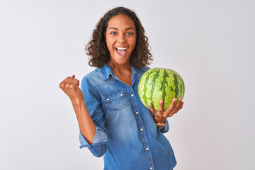 Young brazilian woman holding watermelon standing over isolated white background screaming proud...
