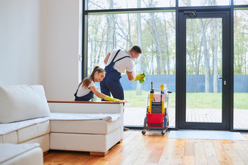 Side view on active job of two caucasian janitors dressed in uniform for cleaning, man clean glass of window, woman wiping dust off sofa. White interior of room, big window