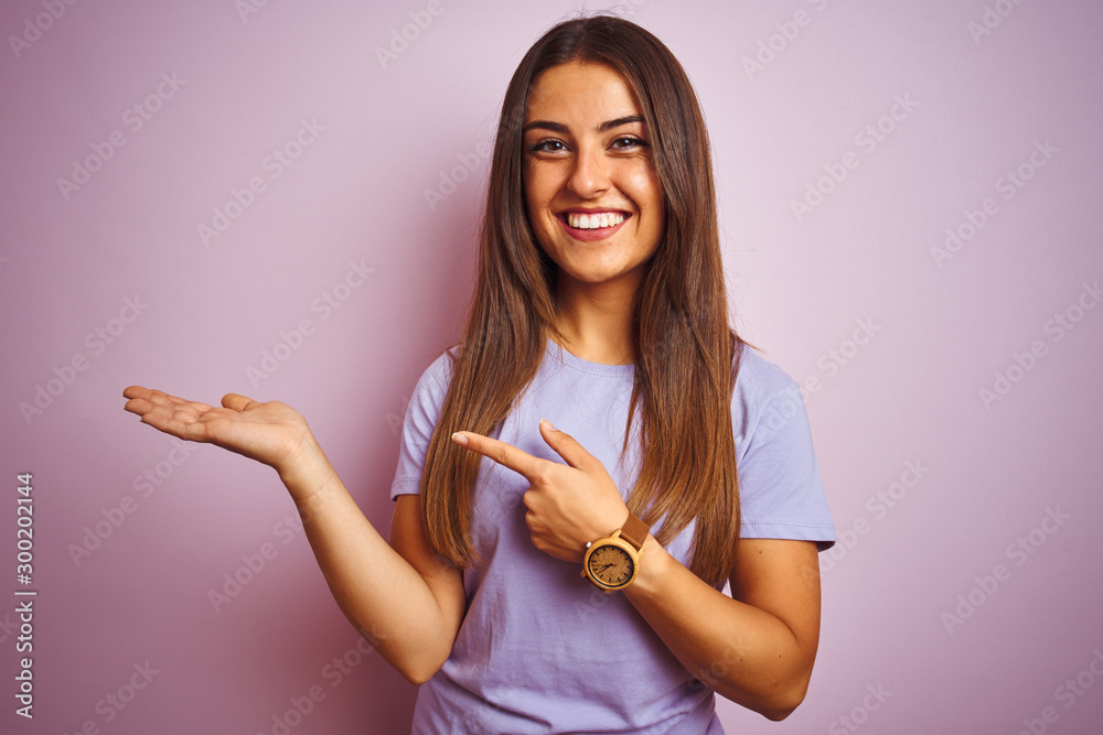 Wall mural Young beautiful woman wearing casual t-shirt standing over isolated pink background amazed and smiling to the camera while presenting with hand and pointing with finger.