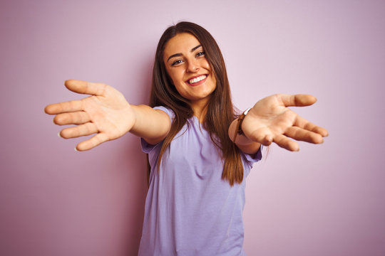 Young beautiful woman wearing casual t-shirt standing over isolated pink background looking at the camera smiling with open arms for hug. Cheerful expression embracing happiness.