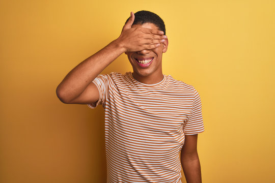 Young handsome arab man wearing striped t-shirt standing over isolated yellow background smiling and laughing with hand on face covering eyes for surprise. Blind concept.