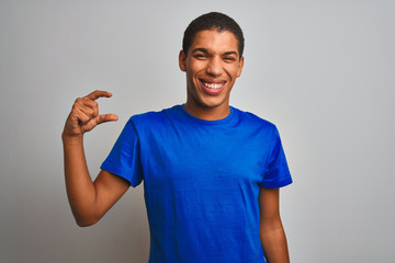 Young handsome arab man wearing blue t-shirt standing over isolated white background smiling and confident gesturing with hand doing small size sign with fingers looking and the camera. Measure