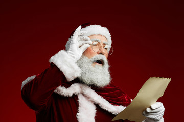 Waist up portrait of classic Santa reading list on parchment standing against red background, copy...