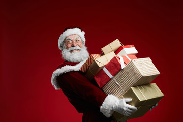 Waist up portrait of smiling Santa Claus holding stack of Christmas presents over red background,...