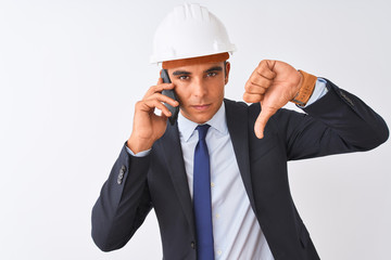 Young architect man wearing helmet talking on smartphone over isolated white background with angry face, negative sign showing dislike with thumbs down, rejection concept
