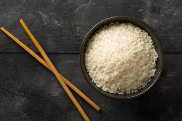 White uncooked, raw long grain rice in black bowl on rustic, dark wooden table top view flat lay...