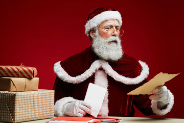 Portrait of classic Santa Claus reading letters sitting at desk with Christmas presents over red background, copy space
