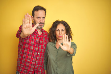 Beautiful middle age couple over isolated yellow background doing stop sing with palm of the hand. Warning expression with negative and serious gesture on the face.